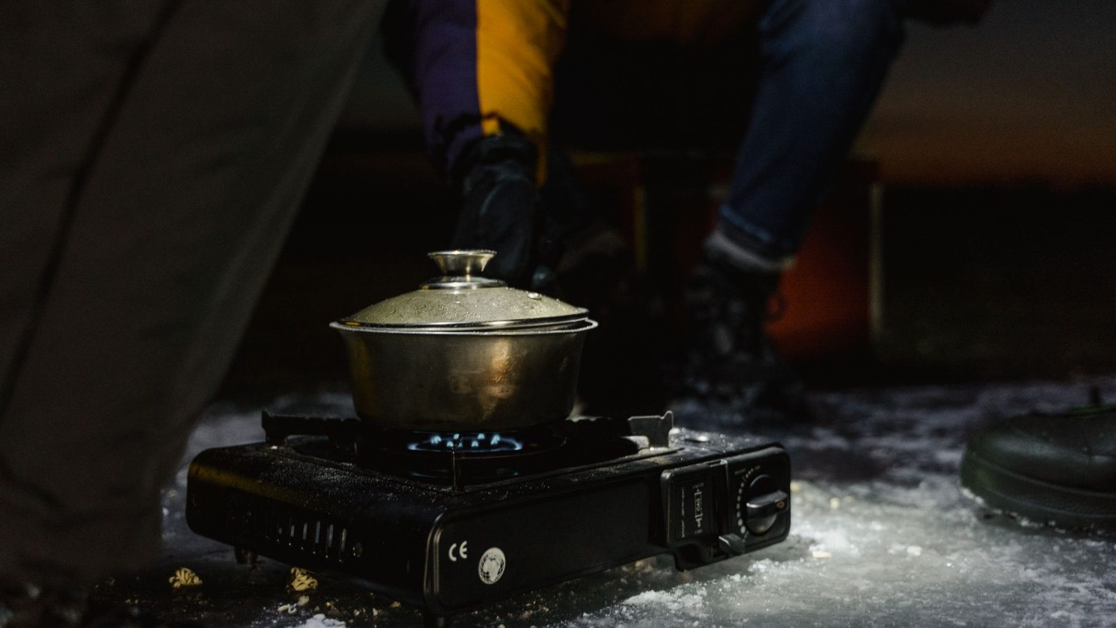 A portable stove with a pot of cooking food, set on snowy ground during winter night camping adventure.