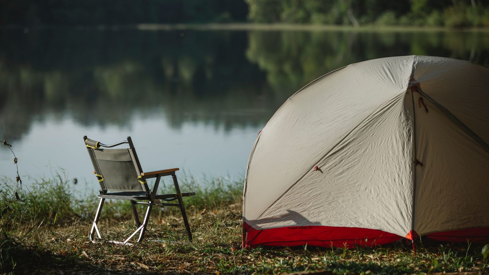 Tent and folding chair on grass coast against trees reflecting in lake in soft sunlight
