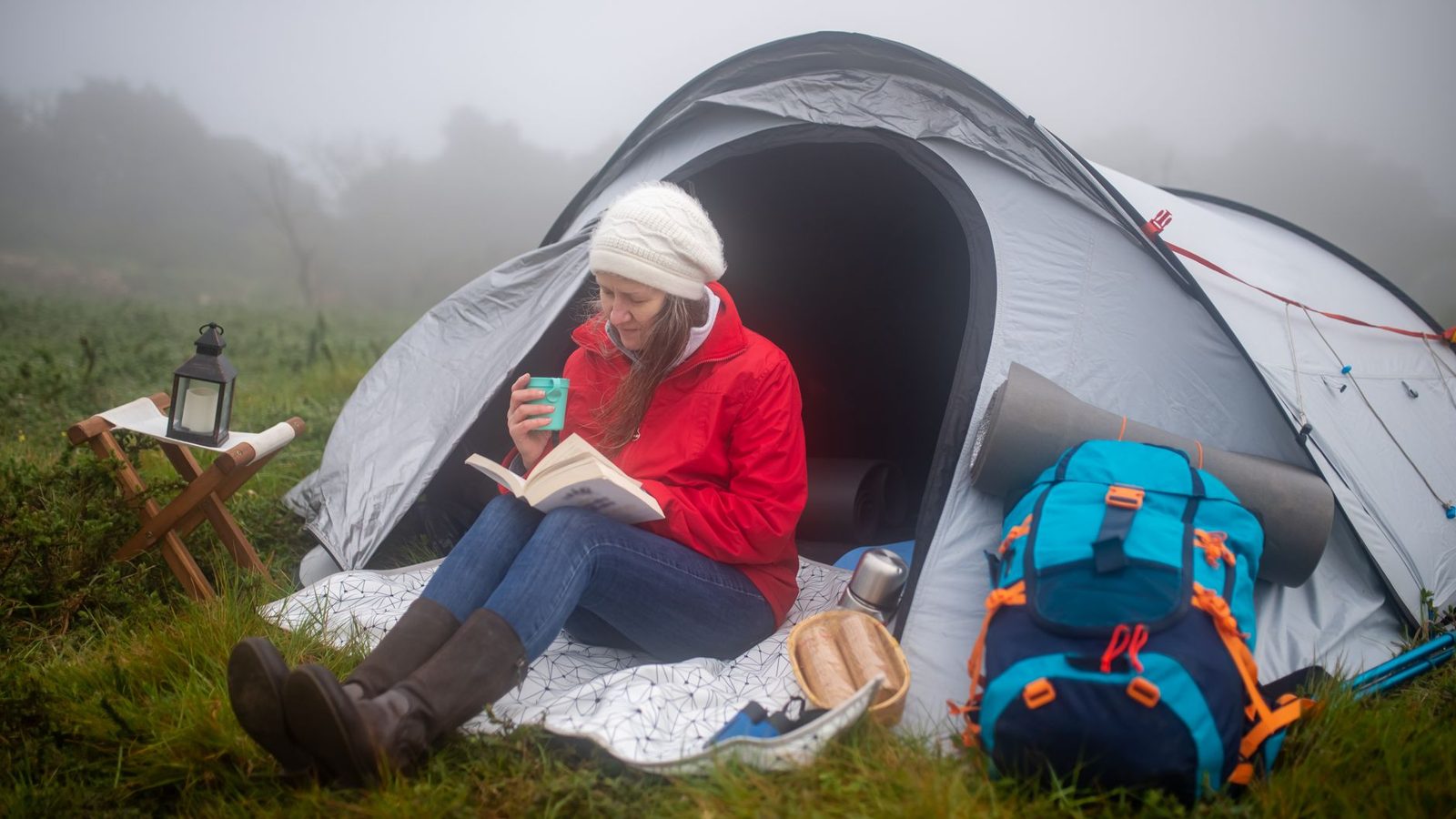 A woman in a red jacket reads a book by her tent on a foggy morning in Portugal.