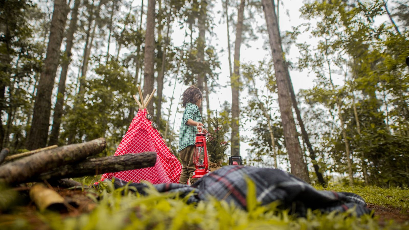 A young child explores a scenic forest campsite with a tent and lantern, surrounded by nature.