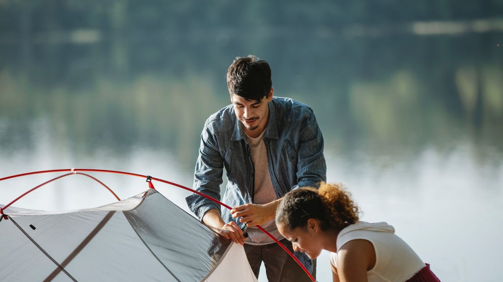 Concentrated young multiracial couple of travelers in casual wear busy with putting up near river