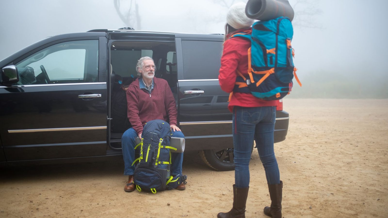An elderly couple with backpacks getting ready for a foggy hiking trip near a vehicle.