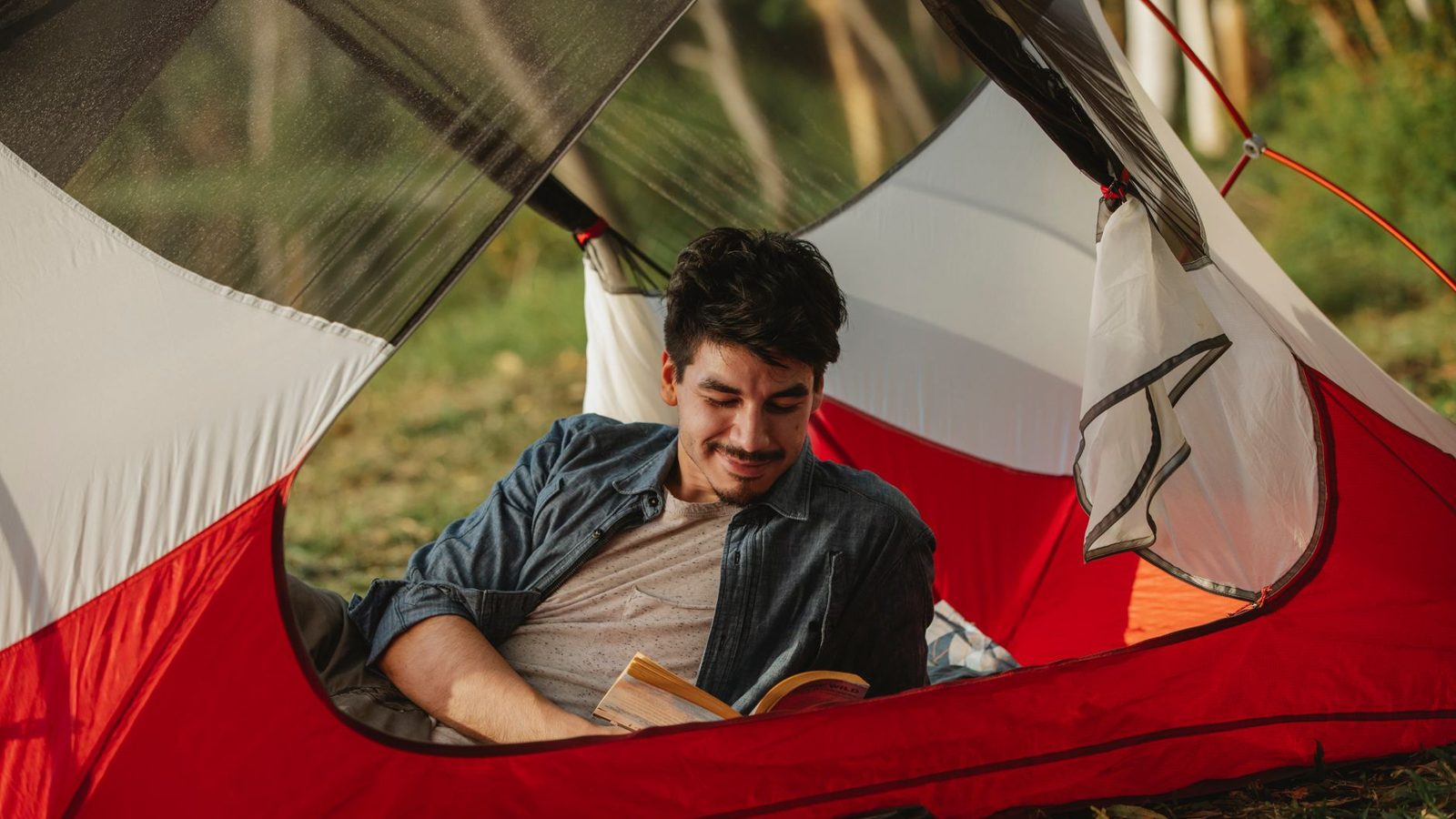 Positive young bearded male traveler smiling and reading interesting book while resting in tent during camping in nature