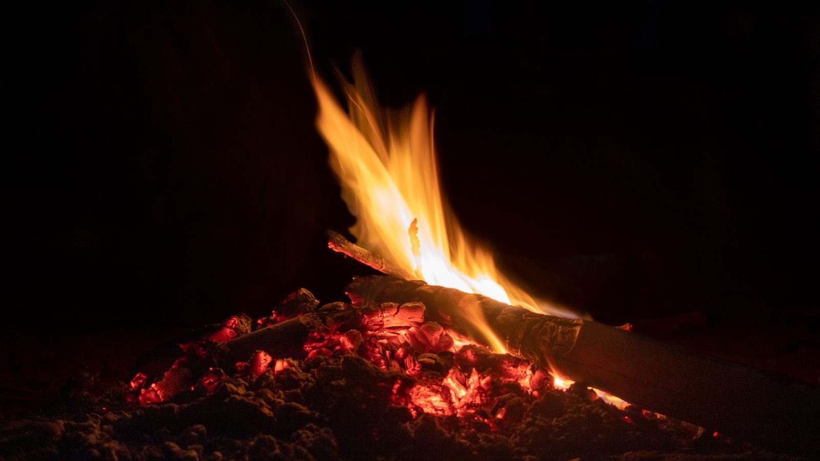 A close-up shot of a campfire with glowing embers, flickering flames set against a dark night backdrop.