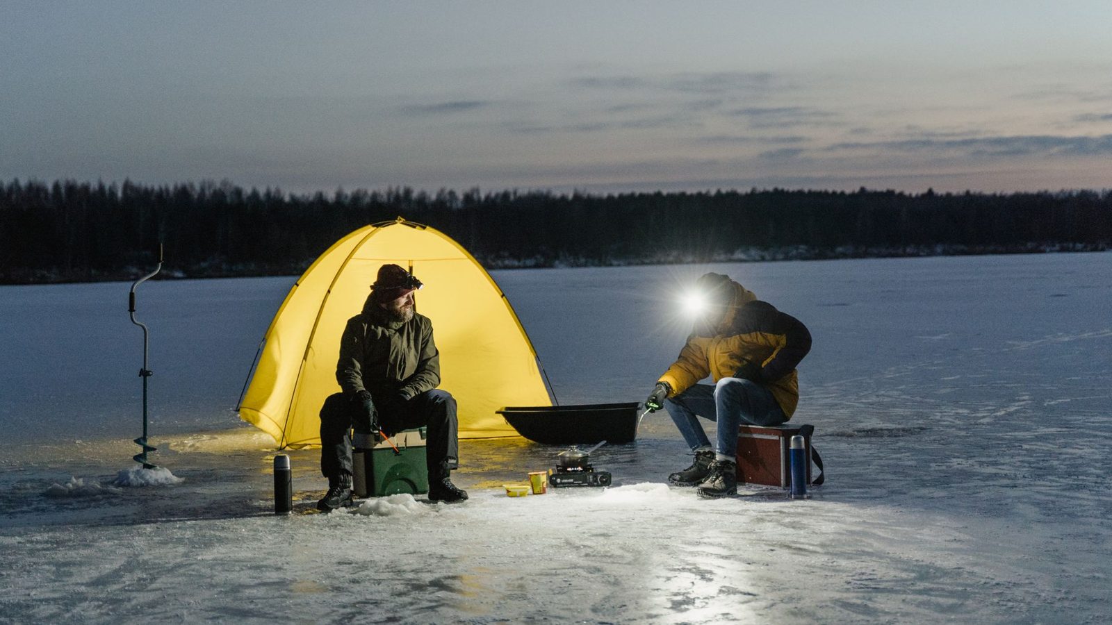 Two adults ice fishing at night near a glowing yellow tent on a frozen lake.