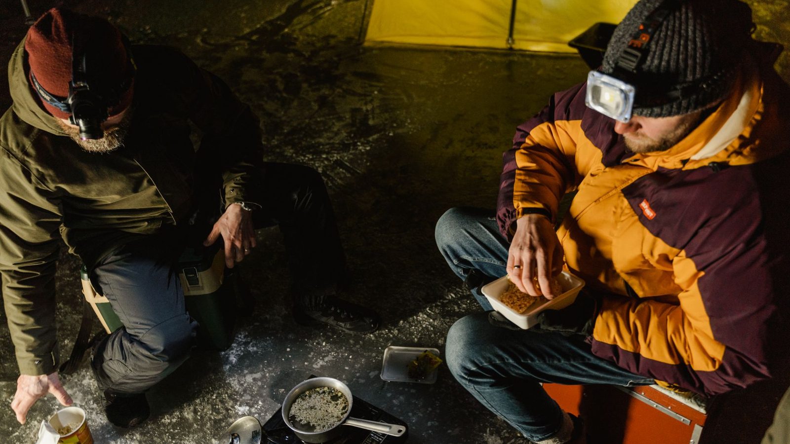 Two men enjoy a warm meal during a winter camping trip at night, sitting beside a tent.
