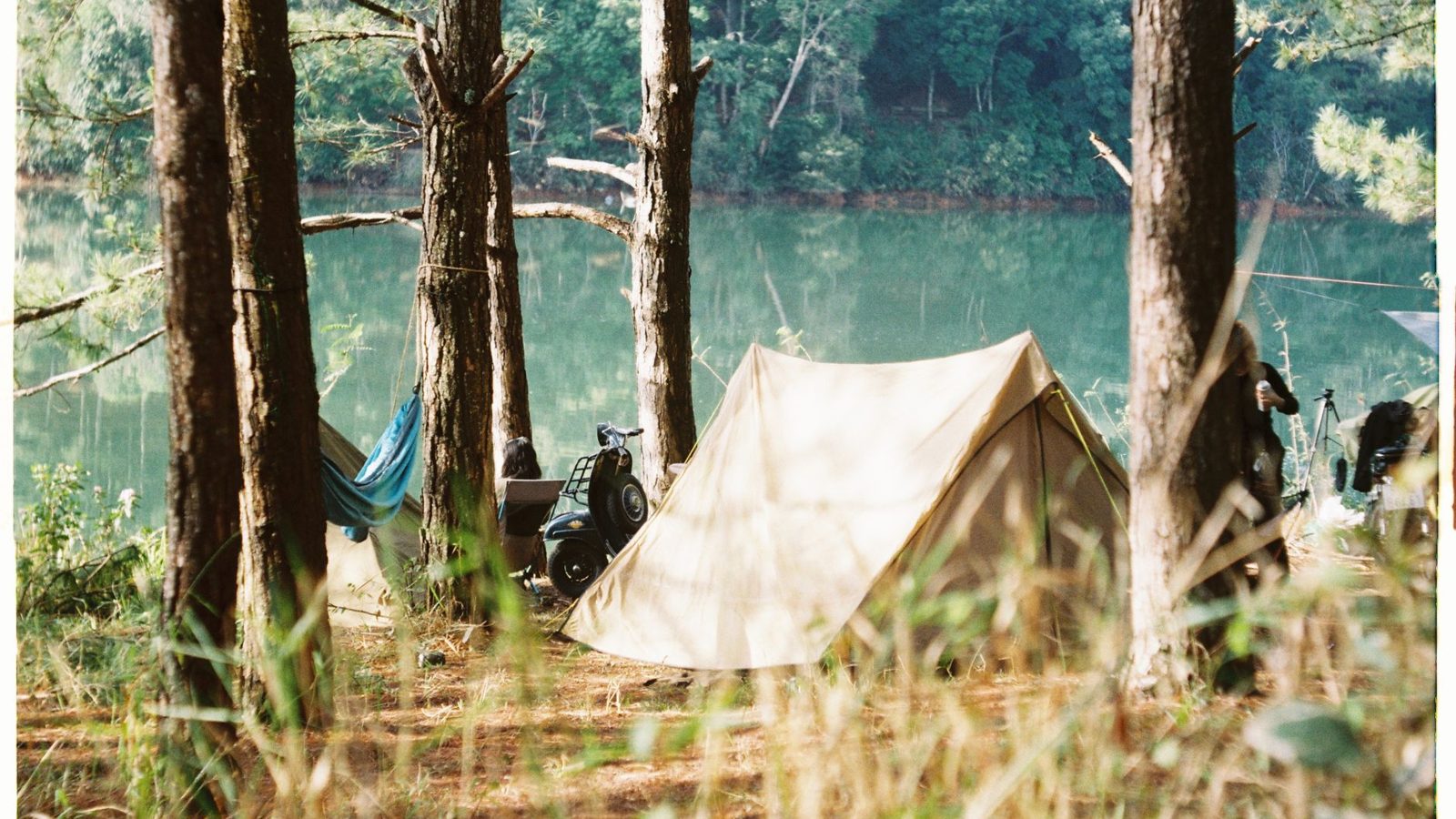 Tranquil camping scene with a tent and hammock in a forest near a lake.