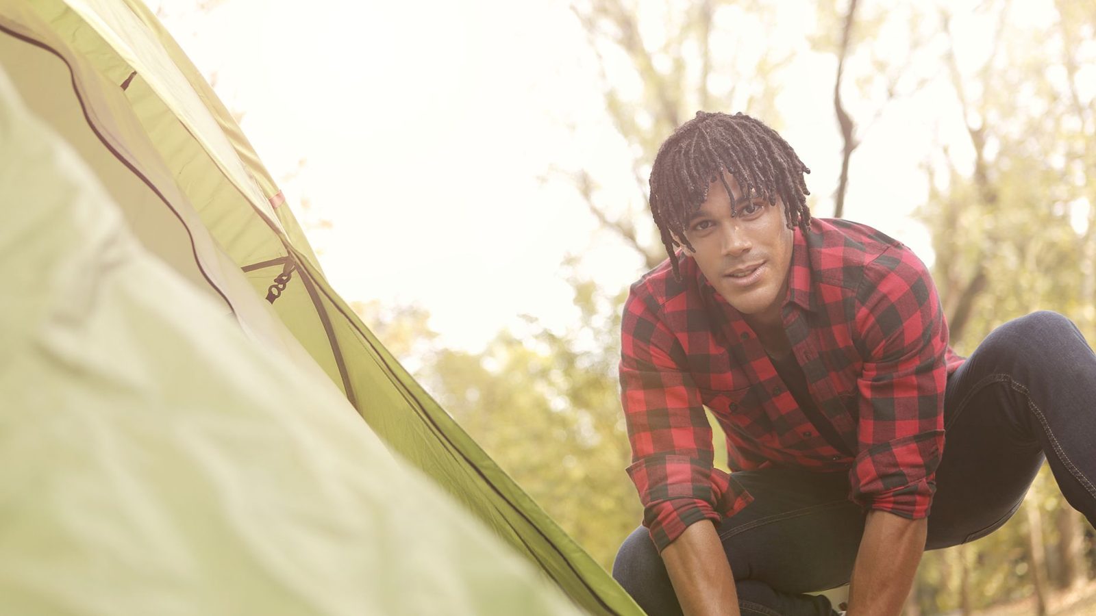 Man in plaid shirt setting up a tent in a sunny forest campsite. Perfect for leisure and adventure themes.